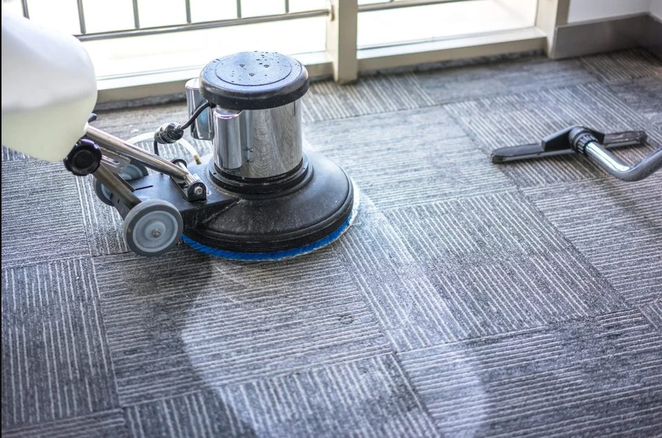 Rotary floor machine cleaning gray commercial carpet tiles beside a window