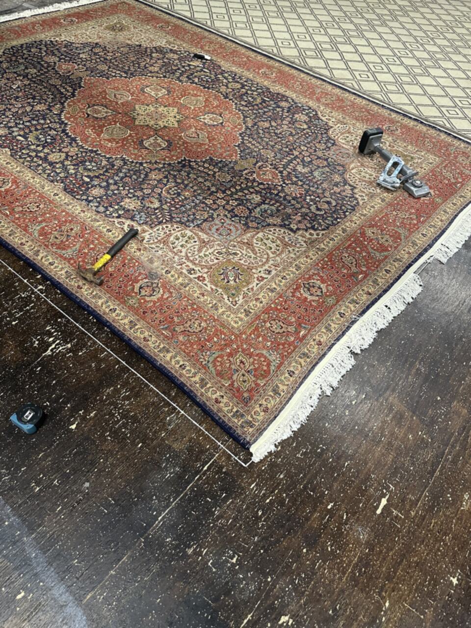 Persian rug being squared on the workshop floor with chalk guide lines, a knee kicker and tape measure during blocking